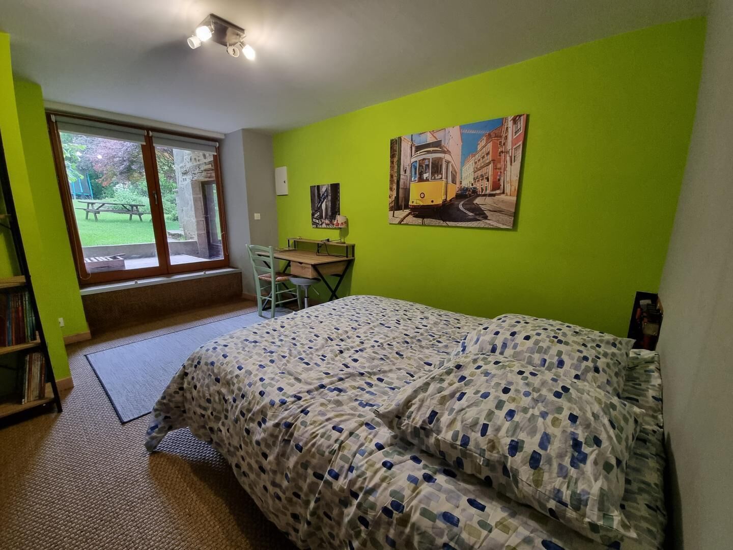 A bedroom with a bed, a desk, and a chair. There is a window with a view of a garden.