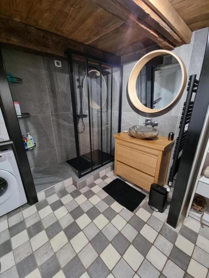 Een badkamer met een zwart-wit geruite vloer, een houten wastafelmeubel, een ronde spiegel en een douchecabine van glas.