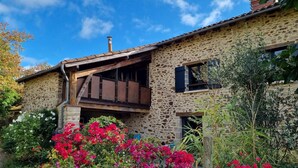 A rustic stone building with a wooden balcony and a garden with vibrant flowers. - Gîtes de France® - Gîte de Larigaldie (SAINT CIRGUES)