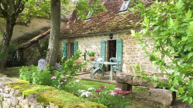 Una casa de piedra con techo de tejas, un muro de piedra y un jardín con flores y un árbol.
