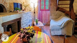 A rustic room with exposed stone walls, a wooden fireplace, a television, a dining table with a colorful table runner, and a cozy sofa. - Gîtes de France® - Le Fournil (SOULOMES)