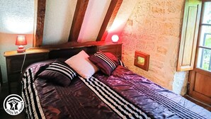 A bedroom with a wooden ceiling, a bed with striped bedding, a nightstand with a lamp, and a small window with a view of greenery. - Gîtes de France® - Le Fournil (SOULOMES)