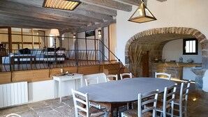 Uma sala de jantar com mesa redonda e cadeiras brancas, piso de pedra e arco de pedra que dá acesso a uma cozinha.