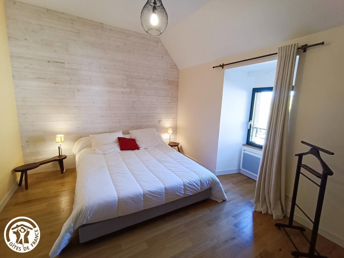 A bedroom with a wooden headboard, a bed with white bedding and a red pillow, two bedside lamps, a window with curtains, and a wooden floor.