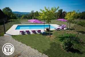 Una piscina con sillas de descanso y sombrillas, rodeada de césped y árboles.