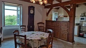 A dining area with a round table covered in a floral tablecloth, wooden chairs, a large wall clock, and a kitchen area with wooden cabinets and a tiled backsplash.