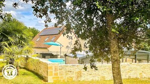 Una casa con piscina, un patio con mobiliario exterior y un árbol en primer plano.