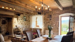A rustic interior with stone walls, a wooden ceiling, a chandelier, a dining table with chairs, and a view through a window. - Gîtes de France® - Le Hameau du Quercy - Stone spirit (FRONTENAC)