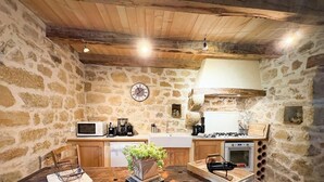 A rustic kitchen with stone walls, wooden ceiling beams, a clock, and basic appliances. - Gîtes de France® - Le Hameau du Quercy - Stone spirit (FRONTENAC)