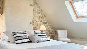 A bedroom with a stone wall, a skylight, a bed with a quilted blanket, and a chair. - Gîtes de France® - Le Hameau du Quercy - Stone spirit (FRONTENAC)