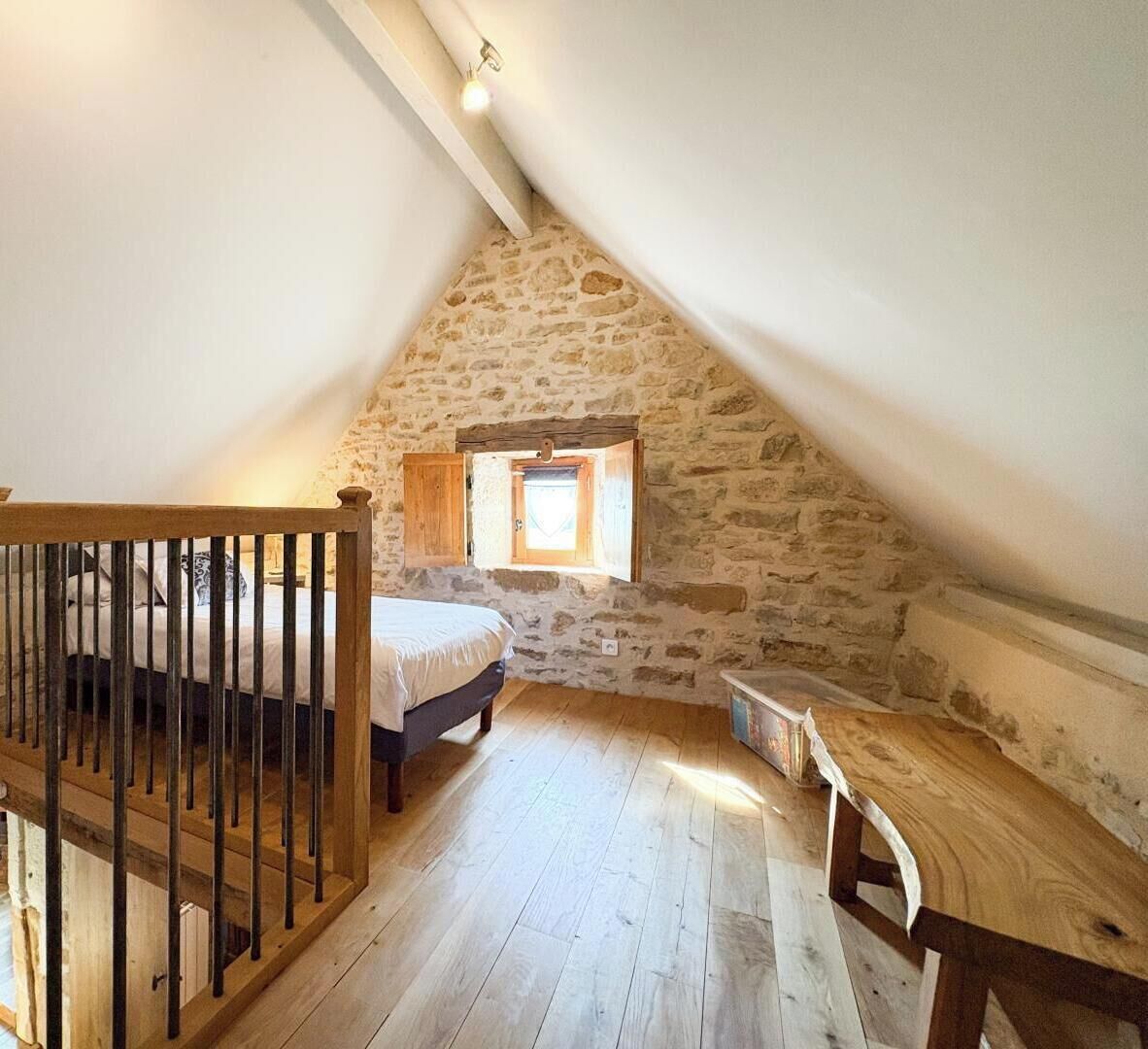 Une chambre à coucher avec un plafond en pente, des murs en pierre, un lit en bois, une petite table et une fenêtre.