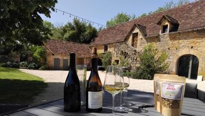 A table with two wine bottles, a wine glass, and a bag of snacks in an outdoor setting with stone buildings in the background. - Gîtes de France® - Gîte du Tilleul (FAYCELLES)