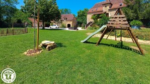 A well-maintained grassy area with a wooden play structure, a stone wall, and a stone building with a tiled roof in the background. - Gîtes de France® - Gîte du Tilleul (FAYCELLES)