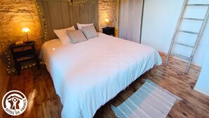 A bedroom with a stone wall, wooden ceiling, a bed with white bedding, two bedside lamps, and a wooden ladder. - Gîtes de France® - Gîte du Tilleul (FAYCELLES)