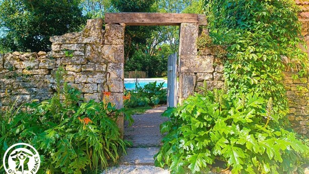 A stone archway leading to a pool area surrounded by lush greenery and a stone wall. - Gîtes de France® - Gîte du Tilleul (FAYCELLES)