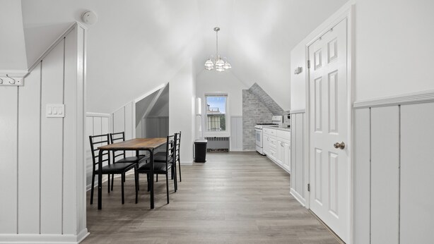 A narrow, white-walled kitchen with a wooden dining table and chairs, a hanging light fixture, and a view to a window. - Cozy Studio with reading nook (White Plains)