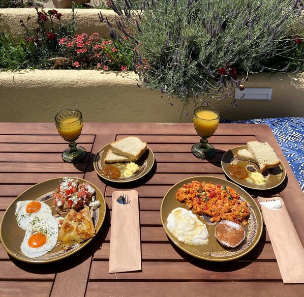 A table with breakfast plates, glasses of orange juice, and a view of a garden with flowers and plants. - Terra Vecchia Suites (Santorini)