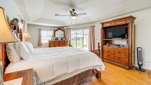 A bedroom with a large bed, wooden headboard, a ceiling fan, a TV on a wooden cabinet, and a standing fan. - Port Charlotte Home ~ 3 Mi to Beach & Harbor! (Port Charlotte)