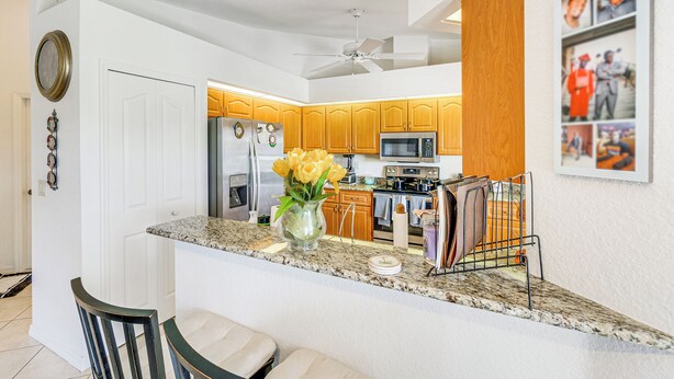 A kitchen with wooden cabinets, granite countertops, a refrigerator, microwave, and a ceiling fan. - Port Charlotte Home ~ 3 Mi to Beach & Harbor! (Port Charlotte)