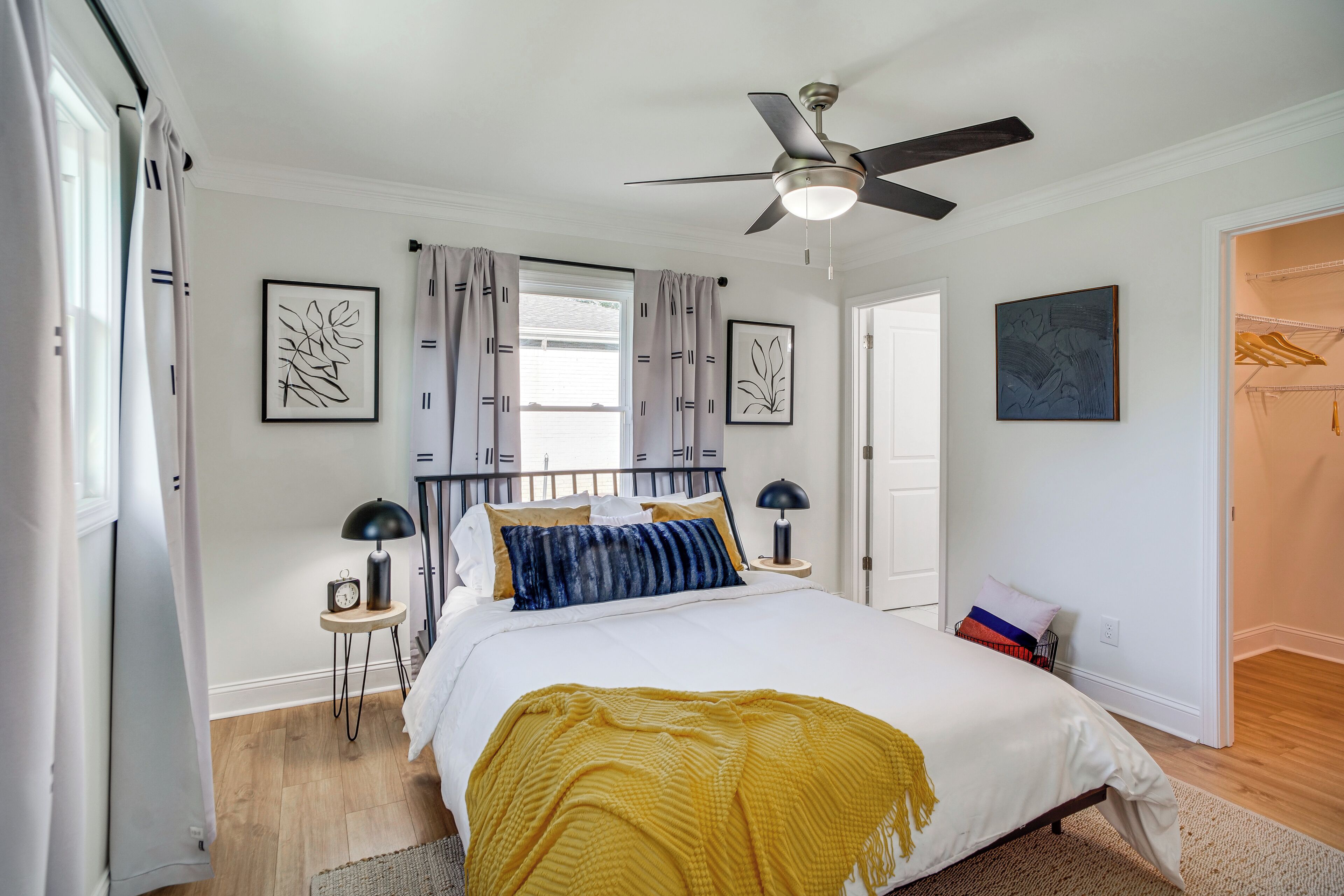 Une chambre à coucher comprenant un lit, un ventilateur de plafond, une fenêtre avec des rideaux, une table de chevet avec une lampe et des œuvres d’art murales.