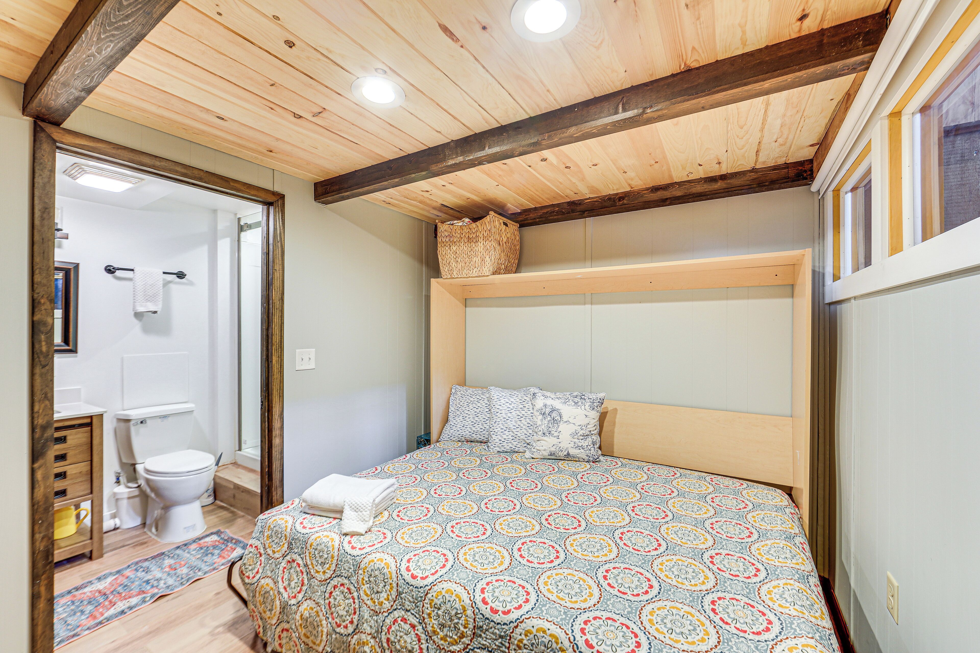 A bedroom with a wooden ceiling, a bed with a patterned bedspread, a bedside table, and a window with a view.
