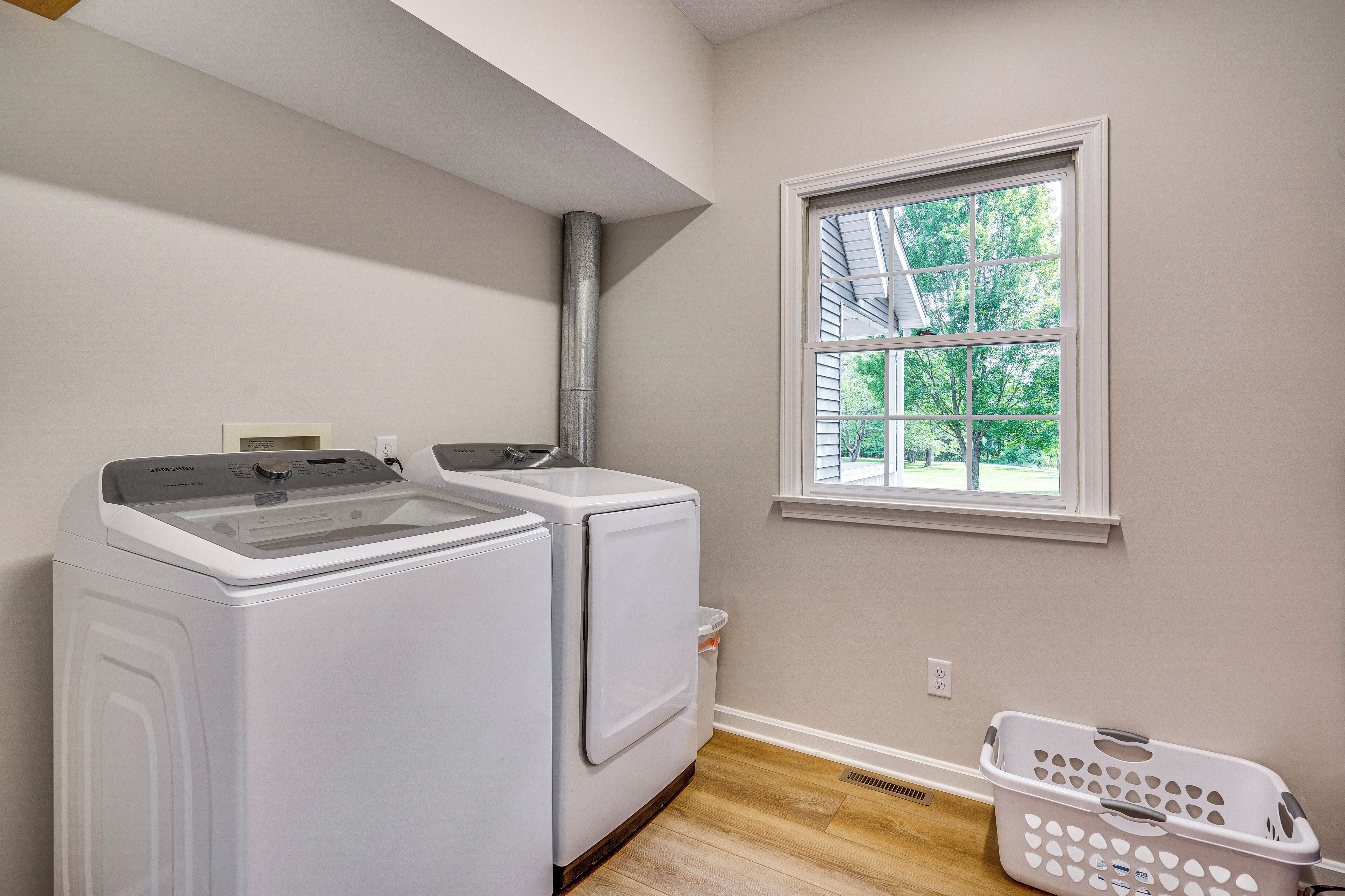 A laundry room with two white washers and dryers, a window with a view of trees, and a laundry basket on the floor.