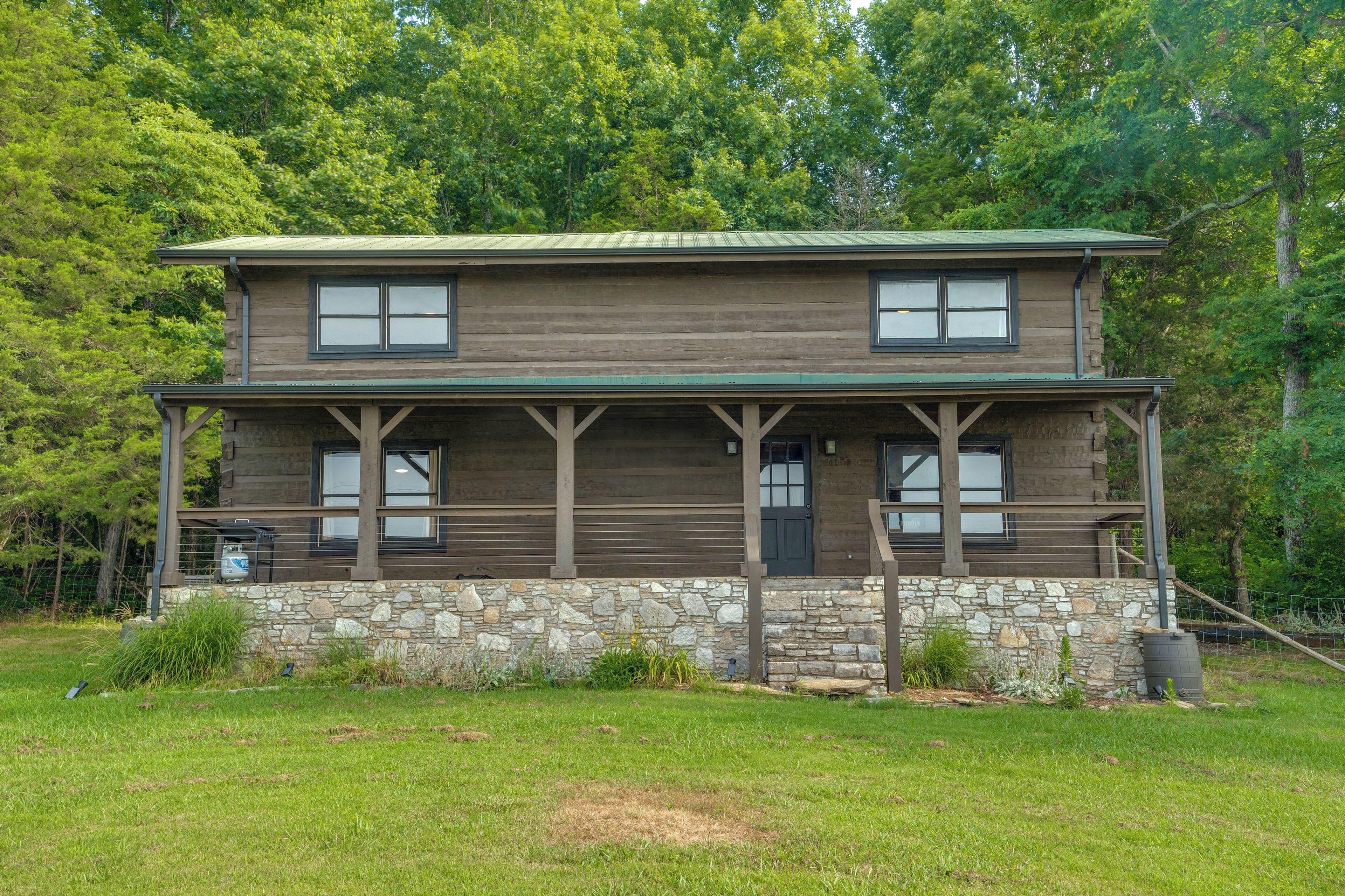 A two-story log cabin with a stone foundation and a green metal roof, surrounded by a grassy yard and dense trees.