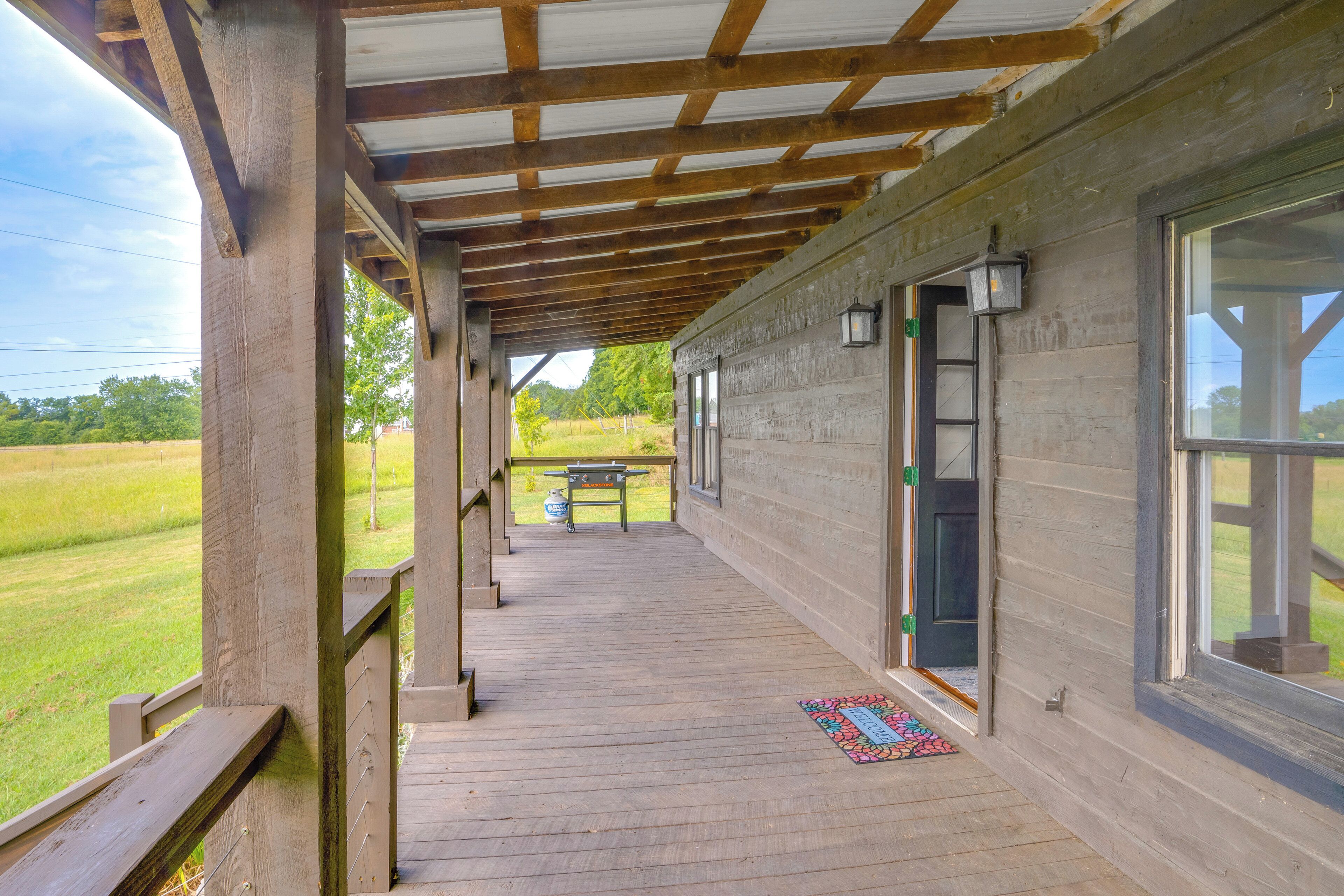 A covered porch with a wooden deck, a bench, and a view of a grassy field.
