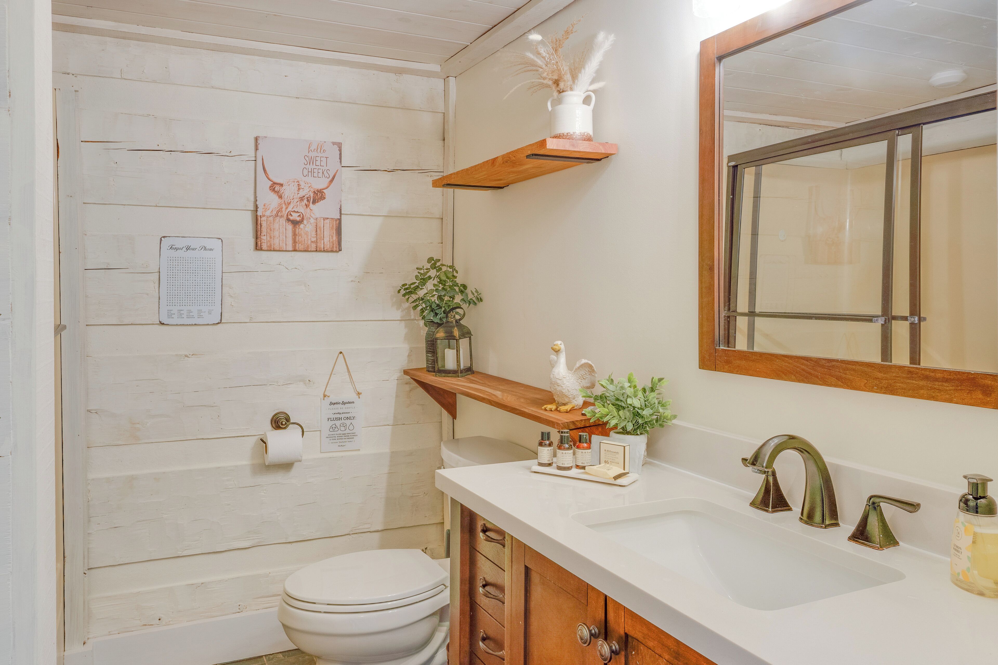 Un baño con un tocador de madera, un lavamanos blanco, un espejo y un inodoro. Hay objetos decorativos en el tocador y un calendario en la pared.