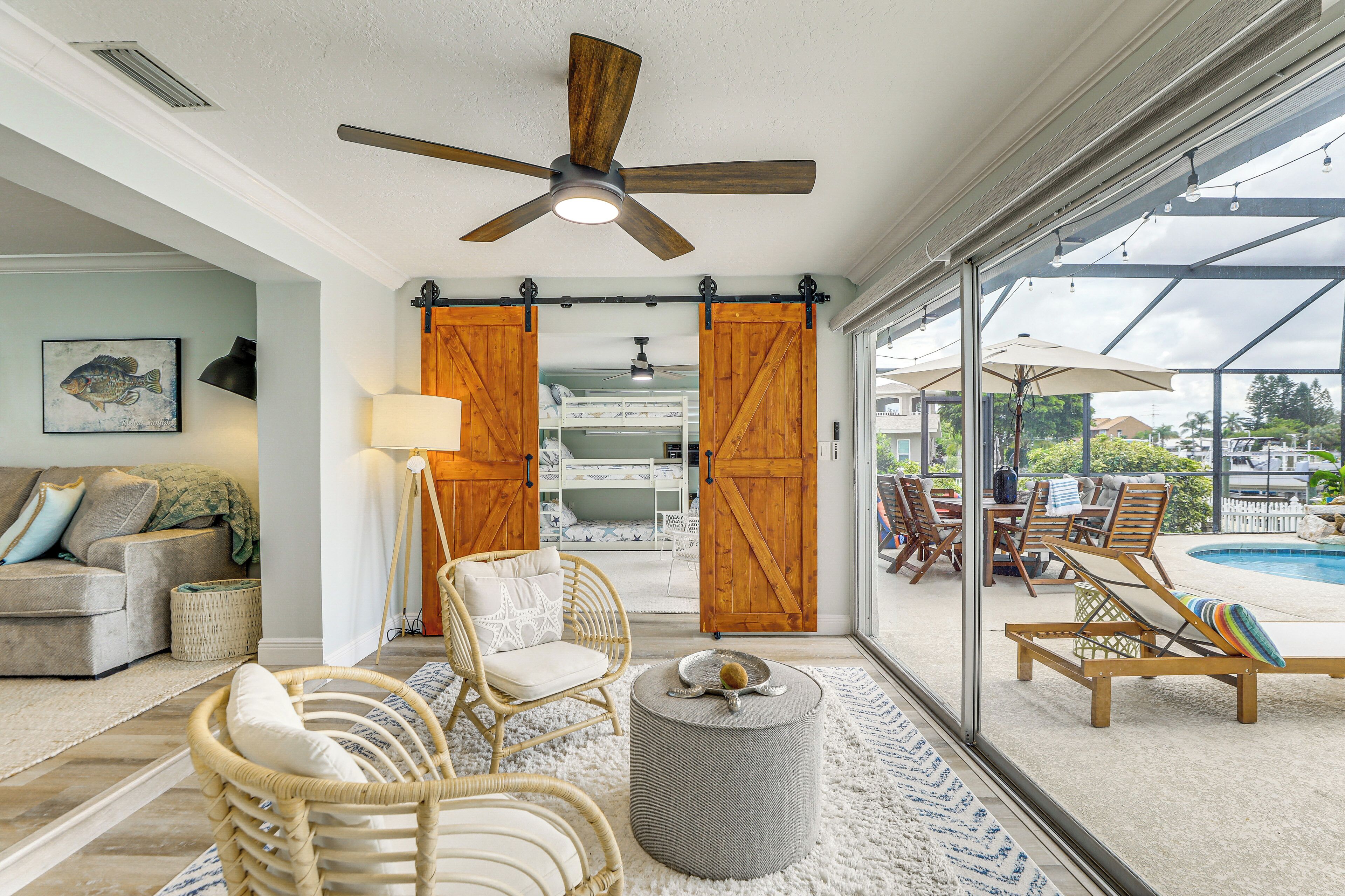 A spacious living area with a ceiling fan, wicker furniture, a round ottoman, and a view of a pool through large glass doors.
