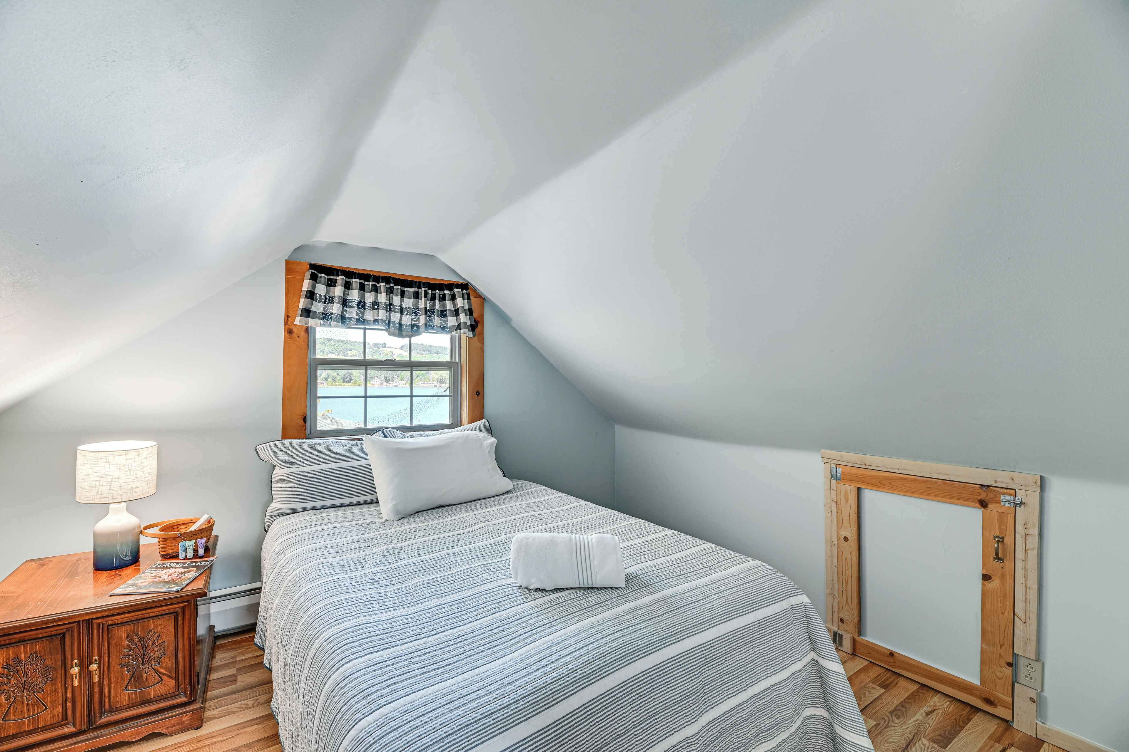 A bedroom with a sloped ceiling, a window with a plaid valance, a bed with striped bedding, a bedside table with a lamp, and a wooden headboard.