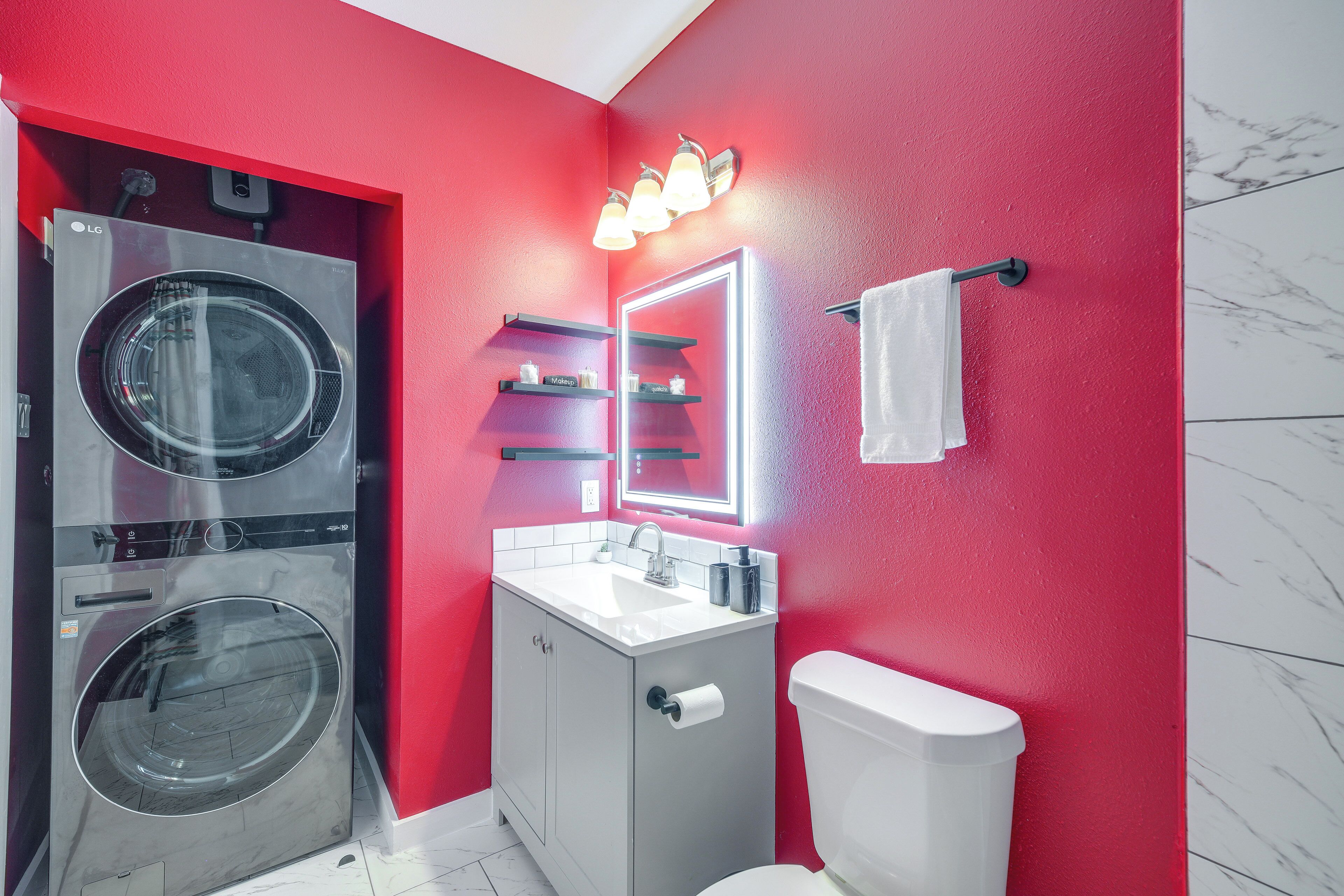 A compact bathroom with a red wall, a white toilet, a sink with a mirror, and a washing machine.