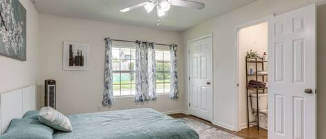 Une chambre à coucher comprenant un lit, un ventilateur de plafond, une fenêtre avec des rideaux et une porte donnant sur une salle de bain.