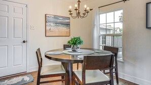 Une salle à manger avec une table ronde, des chaises en bois, un lustre, un tableau représentant une plage et une fenêtre donnant sur des maisons.