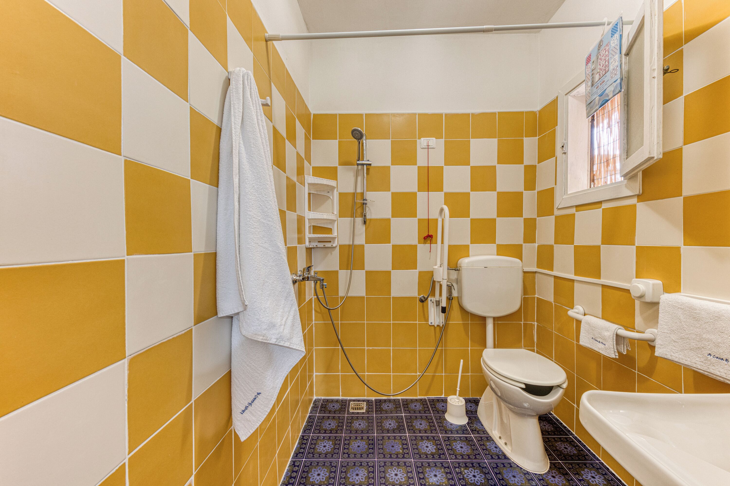 Ein Badezimmer mit gelb-weißen Schachbrettfliesen, einer weißen Toilette, einer Dusche inklusive Handbrause und einer Badewanne.
