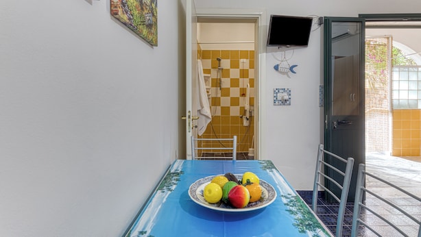 Ruokailutila, jossa on kahdelle katettu pöytä, hedelmävati, seinälle asennettu televisio ja ovi, joka johtaa kylpyhuoneeseen, jossa on ruutukuvioinen lattia.