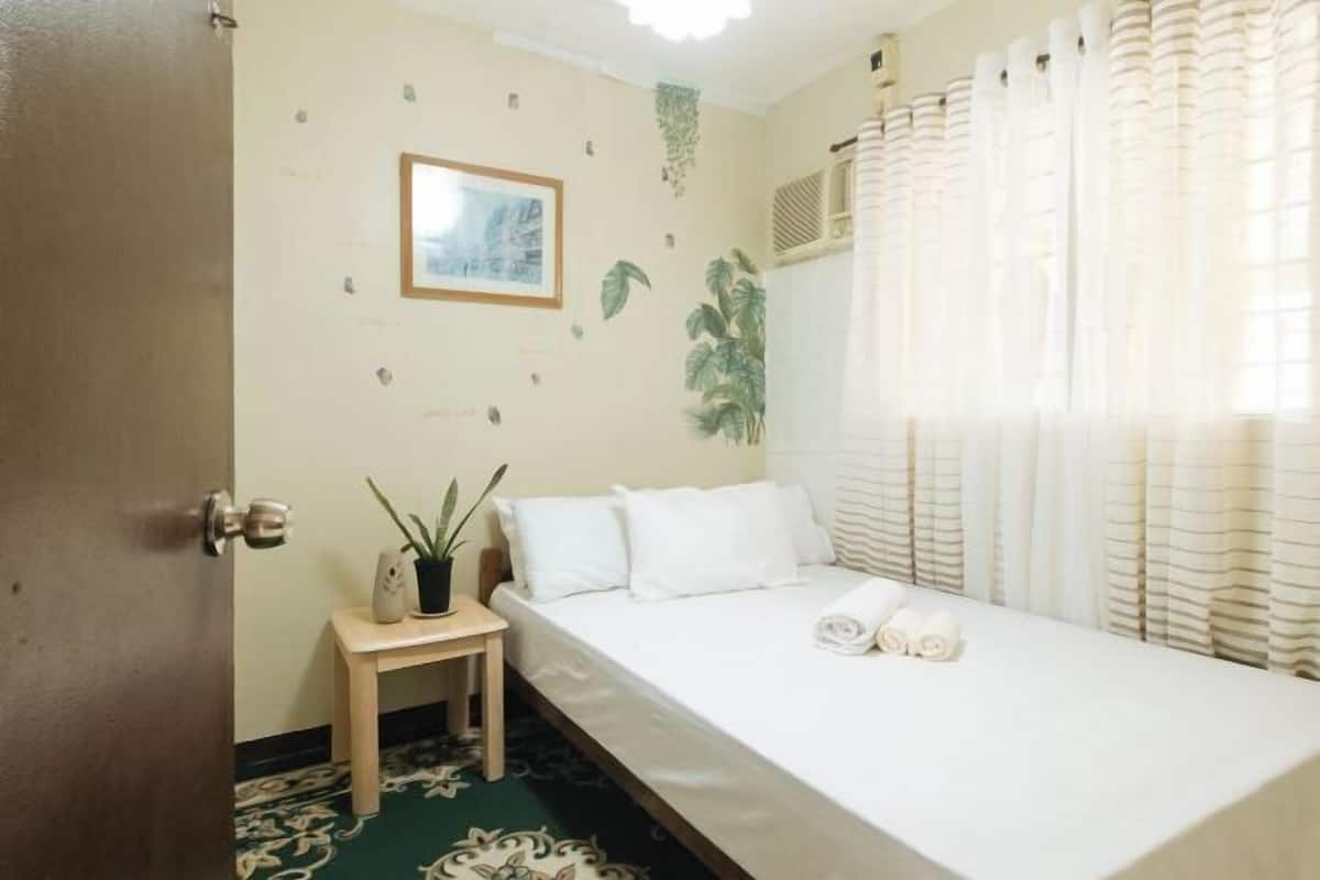 Una cama individual con ropa blanca, una mesita de noche con una planta, un cuadro en la pared y una ventana con cortinas.
