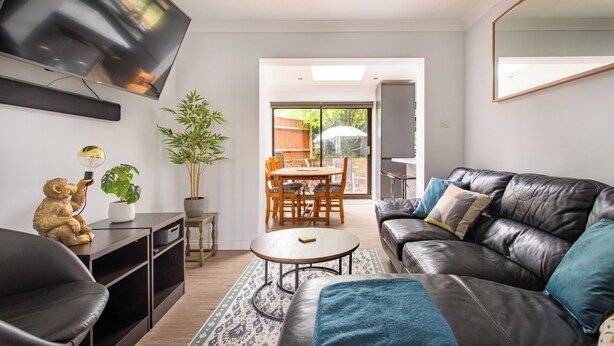 A modern living room with a leather sofa, a flat-screen TV mounted on the wall, a dining table with chairs, and a large window with a view of the outdoors. - Charming Retreat Near Grange Hill (Central Line) (Greater London)