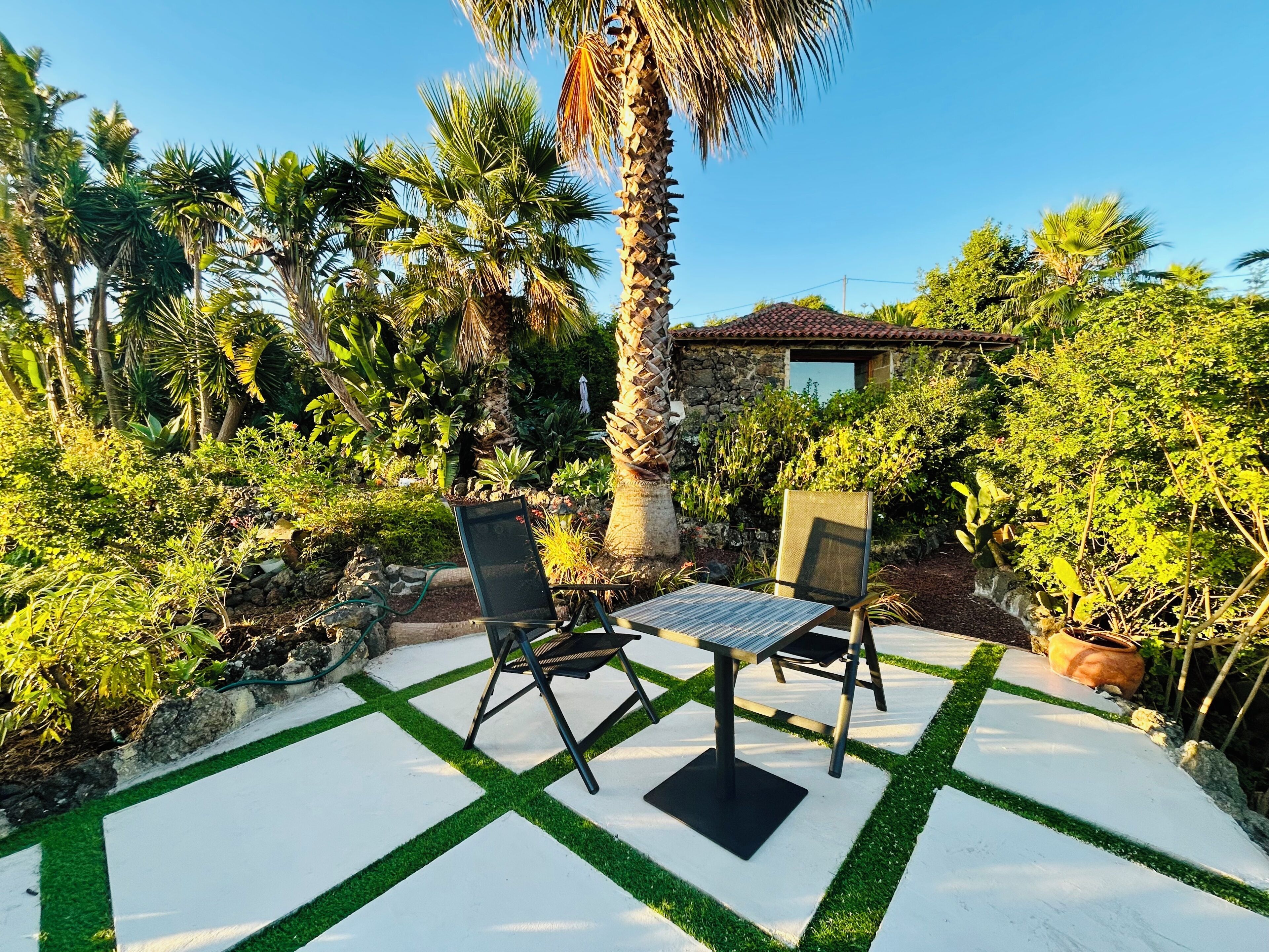 An outdoor patio with two chairs and a table, surrounded by lush greenery and a stone wall.