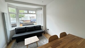 A modern living room with a black sofa, a wooden coffee table, and a dining table with chairs. Large windows provide a view of the street outside. - Newly Renovated 4BD Home, 11 Mins to ExCel London! (London)