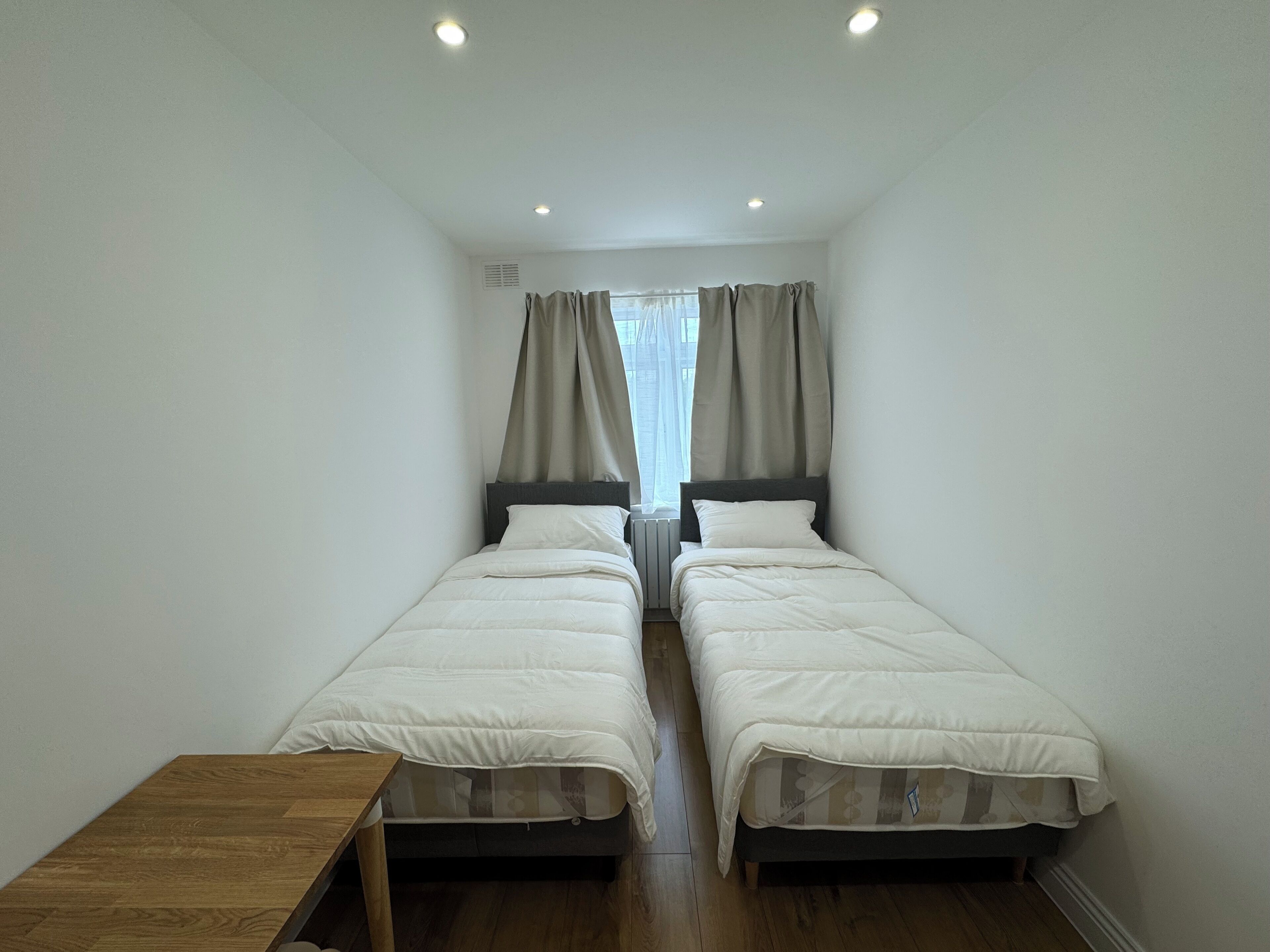 A small, well-lit room with two single beds, a wooden table, and a window with curtains.
