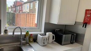 A compact kitchen with a stainless steel sink, a window overlooking a residential area, a washing machine, a toaster, and a kettle. - Newly Renovated 4BD Home, 11 Mins to ExCel London! (London)