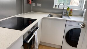 A compact kitchen with a built-in stove, a dishwasher, and a window with a view of buildings. - Newly Renovated 4BD Home, 11 Mins to ExCel London! (London)