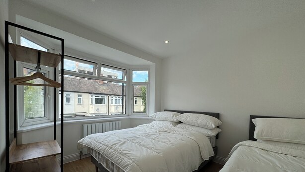 A bedroom with a bed, a wooden chair, a radiator, and a window with a view of buildings. - Newly Renovated 4BD Home, 11 Mins to ExCel London! (London)