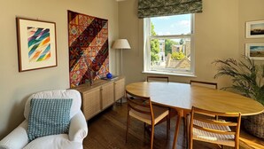A dining area with a wooden table and chairs, a white cushioned chair, a lamp, and artwork on the walls. - Hidden Gem w/Private Terrace, Fulham Broadway! (London)