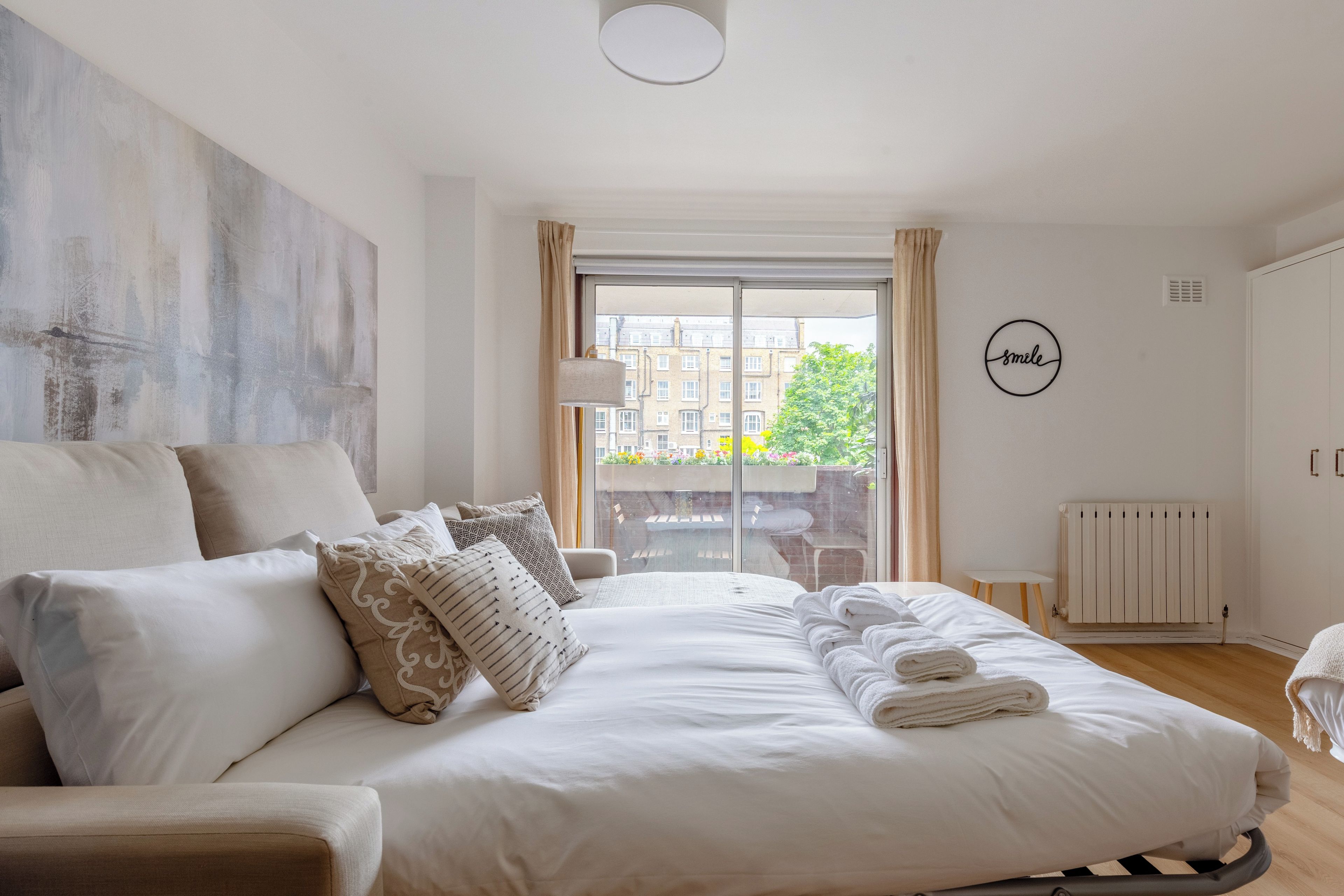 A modern bedroom with a large bed, a view of buildings through the window, and a 'smile' wall decoration.