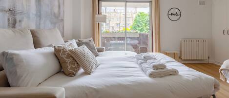 A modern bedroom with a large bed, a view of buildings through the window, and a 'smile' wall decoration.