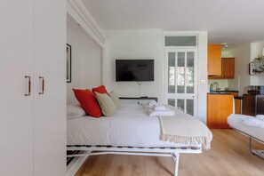 A bedroom with a bed, a TV, a bathroom with a mirror, and a kitchen area. - Cosy Refurbished Studio w Pvt Balcony Kensington (London)