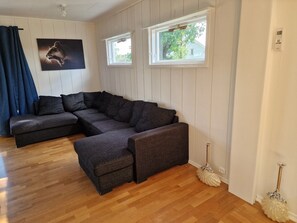 Een moderne woonkamer met een hoekbank, houten vloeren en twee ramen met zonwering.