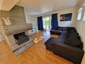 Een ruime woonkamer met een stenen openhaard, een donkerbruine zithoek en een groot raam met gordijnen.